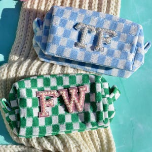 May include: Three rectangular zippered pouches featuring a checkered pattern in pink, light blue, and green. Each pouch displays two large capital letters embellished with white faux pearls. The pink pouch has AB, the blue has TF, and the green has FW.