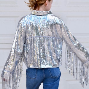 Puede incluir: Chaqueta plateada con lentejuelas y mangas con flecos. La palabra "MRS" está bordada en la parte posterior del cuello. La chaqueta se combina con vaqueros azules. Una pieza elegante y llamativa para ocasiones especiales.