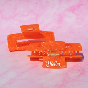Puede incluir: Dos pinzas para el pelo naranjas con efecto marmolado. Una pinza tiene el nombre "Shelby" impreso en blanco. Las pinzas son de plástico duro brillante y tienen bisagras metálicas doradas. El fondo es un patrón marmolado rosa y blanco.