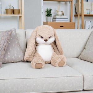 Könnte beinhalten: Ein großes Plüschkaninchen mit wütendem Gesichtsausdruck sitzt auf einem hellen Sofa. Das Kaninchen ist hauptsächlich hellbraun mit einem weißen Bauch und Innenohren. Es hat lange, schlaffe Ohren und ein strenges Gesicht mit roten Markierungen über den Augen.