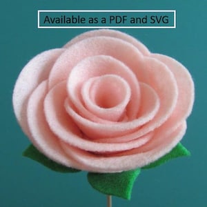 May include: A pale pink felt rose with green leaves and a wooden stem. The flower has multiple layers of petals and the text "Available as a PDF and SVG" is in a white box at the top.