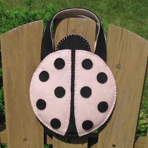 Puede incluir: Un bolso hecho a mano con forma de mariquita, hecho de fieltro rosa y negro. El bolso presenta un cuerpo redondeado con lunares negros y un asa negra. El bolso está sobre una valla de madera.