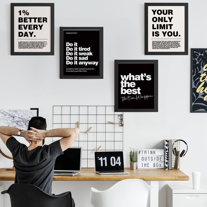 May include: A workspace featuring framed motivational posters with black frames and white text. The posters display phrases like "1% Better Every Day" and "Your Only Limit Is You." A laptop shows the time "11 04."