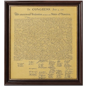 May include: A framed replica of the Declaration of Independence featuring black printed text on aged, tan parchment paper. The document is enclosed in a dark brown wooden frame with a thin gold inner border, displaying the historic text and signatures.
