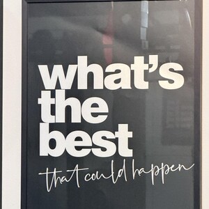 May include: Black framed print with white text that reads "what's the best that could happen" in a mix of bold and script fonts. The print is mounted on a white wall.