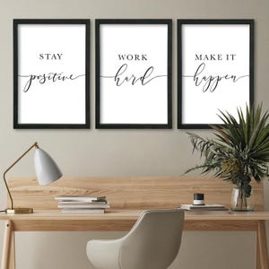 May include: Three framed wall art prints with motivational quotes. The prints are in black frames with white backgrounds. The first print says "Stay Positive", the second says "Work Hard", and the third says "Make it Happen". The prints are displayed on a wooden desk.