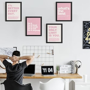 May include: A workspace with motivational framed prints in black frames and pink backgrounds. The prints feature phrases like "1% BETTER EVERY DAY" and "YOUR ONLY LIMIT IS YOU." A person sits at a wooden desk with a laptop, a digital clock displaying "11 04", and a sign that says "#THINK OUTSIDE THE BOX."