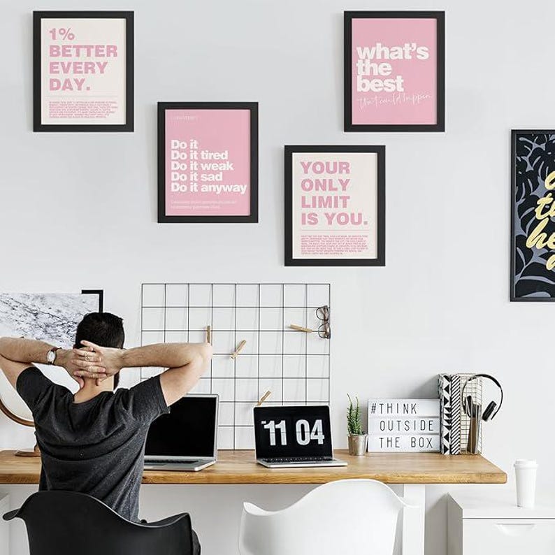 May include: A workspace with motivational framed prints in black frames and pink backgrounds. The prints feature phrases like "1% BETTER EVERY DAY" and "YOUR ONLY LIMIT IS YOU." A person sits at a wooden desk with a laptop, a digital clock displaying "11 04", and a sign that says "#THINK OUTSIDE THE BOX."
