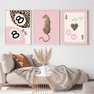 May include: A set of three framed wall art prints. The left print features two billiard balls with leopard print and the word Lucky. The center print shows a leopard on a pink background. The right print displays an ace card with a leopard heart and lips.
