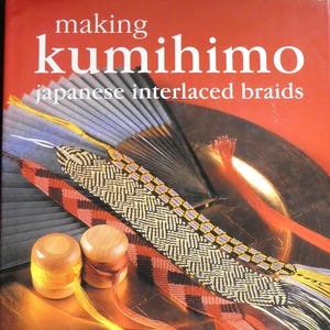 以下が含まれることがあります： Rodrick Owen著の書籍「making kumihimo japanese interlaced braids」。赤い背景に金色の皿、灰色の扇子、黄色と茶色の糸が巻かれた2つの木製スプール、そして複雑に編み込まれた組紐が表紙にデザインされています。