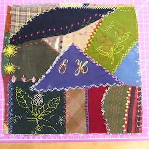 May include: A patchwork textile with various fabric pieces in shades of green, blue, red, and brown. The textile features embroidered floral and geometric designs, with the letters "E K" embroidered on a blue triangle. The edges are stitched with contrasting thread.