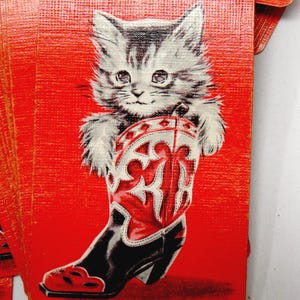 May include: A stack of red playing cards featuring a kitten holding a red, white, and black cowboy boot. The kitten has gray and white fur. The cards have a textured surface.