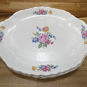 May include: White oval serving platter with a scalloped edge and floral design. The platter features pink, yellow, and blue flowers with green leaves. The rim has a silver trim. The platter is on a wooden surface.