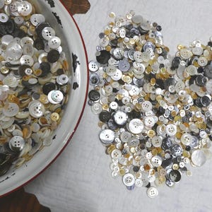 May include: A large collection of assorted sewing buttons in shades of white, cream, gray, and black. Many buttons are arranged in a heart shape on a white surface, while others are contained within a white bowl with a red rim edge.