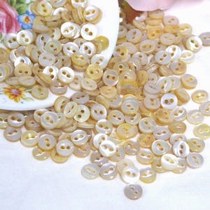 May include: A large collection of small, round, two-hole buttons made of iridescent, cream-colored shell material. The buttons are scattered across a white surface, with many spilling out from a white ceramic bowl decorated with floral patterns.
