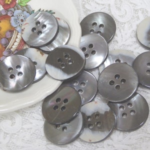 May include: A collection of 12 gray and white mother of pearl buttons with four holes each. The buttons are arranged in a pile on a white lace tablecloth.