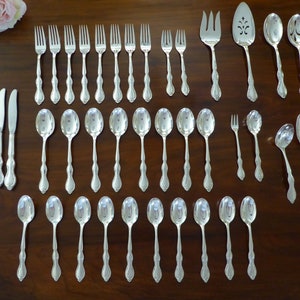 May include: A complete set of silver-colored flatware arranged on a dark wooden surface. The set includes knives, forks, spoons, and serving utensils. The silverware has a decorative handle design. A pink rose is visible in the upper left corner.