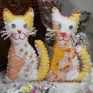 May include: Two handmade fabric cat figurines crafted from patchwork pieces in shades of yellow, white, and peach. Each cat features button eyes, embroidered orange noses and mouths, and white string whiskers with visible blanket stitching.
