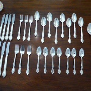 May include: A collection of silver-colored flatware, including knives, forks, and spoons, arranged on a dark wooden surface. The silverware has decorative handles and is likely part of a formal dining set. A single pink rose is visible in the upper left corner.