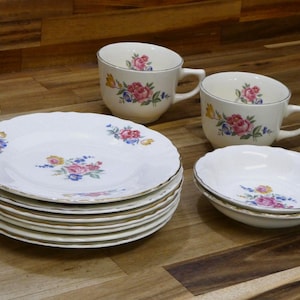 May include: A vintage tea set featuring white porcelain plates, cups, and a bowl, all adorned with floral patterns in pink, blue, and yellow. The set includes a stack of plates, two cups with handles, and a small bowl, all with gold trim.