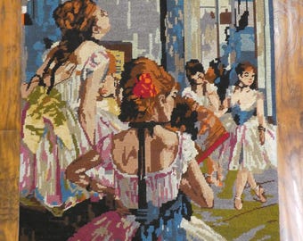 May include: A vintage needlepoint wall hanging depicting a group of ballerinas in a dance studio. The ballerinas are wearing colorful tutus and are in various poses. The background is a muted blue and gray with a window in the back.