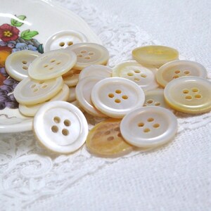 May include: A collection of round, off-white, four-hole buttons with a smooth, pearlescent finish. The buttons vary slightly in shade, ranging from creamy white to light amber, and are scattered in a pile, showcasing their circular shape.