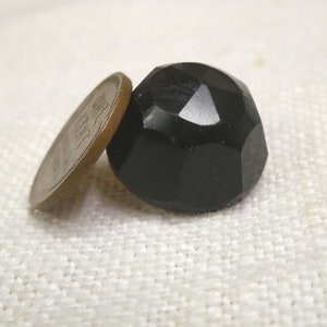 May include: A faceted, black, round bead is positioned next to a penny. The bead has multiple flat surfaces that reflect light. The penny is bronze-colored and partially obscures the bead. The background is a textured, off-white fabric.