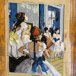 May include: A needlepoint canvas depicting a painting of a dance class by Edgar Degas. The canvas features a group of young women in white tutus, with one woman in the foreground wearing a blue ribbon. The text "Leçon de danse (detail)" is printed at the top of the canvas.