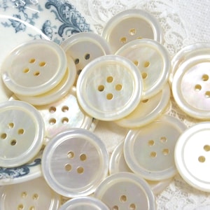 May include: A collection of round, off-white mother-of-pearl buttons, each featuring four central holes and a smooth, iridescent surface. These circular fasteners are arranged in a cluster, displaying their natural pearlescent sheen and texture.