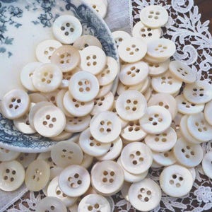 May include: A large collection of round, off-white, four-hole buttons made of mother-of-pearl. The buttons are scattered across a white lace fabric, with many contained within a shallow, white ceramic bowl featuring a blue floral pattern.
