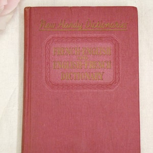 May include: A red hardcover book with gold lettering on the cover. The title is "New Handy Dictionaries French-English and English-French Dictionary".