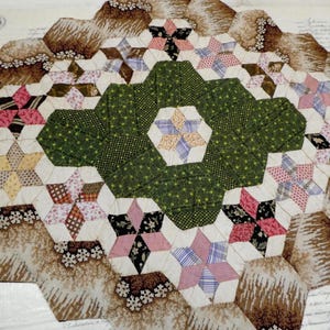 May include: A handmade quilt with a repeating pattern of hexagonal shapes. The quilt features a central green hexagon surrounded by star-shaped patches in various colors, including pink, brown, and white. The quilt's edges are a light brown color.