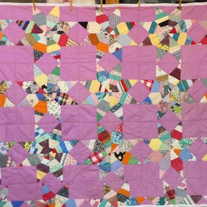 May include: A rectangular quilt featuring a repeating pattern of solid lavender square blocks interspersed with multi-colored patchwork star motifs. The patchwork sections consist of various small fabric scraps in diverse patterns, including plaids, stripes, and polka dots, all framed by a white border.