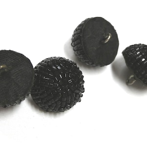 May include: Four black buttons with a beaded design. The buttons have a round shape and a shank on the back for sewing. The buttons are made of a dark fabric and are suitable for various sewing projects.