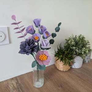 Peut inclure: Un vase en verre transparent contient un bouquet de fleurs en feutre violet, lavande et rose, avec des touches de vert. L'arrangement comprend divers types de fleurs et de feuillage. Le vase est posé sur une surface en bois.
