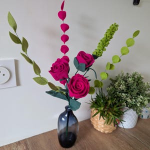 Peut inclure: Un arrangement floral en feutre dans un vase en verre bleu foncé. L'arrangement comprend trois roses roses vibrantes, des feuilles vertes et des tiges avec des accents roses en forme de cœur. De la verdure supplémentaire et de petites plantes en pot sont en arrière-plan.