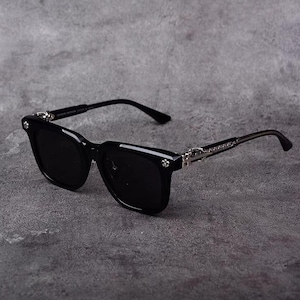 以下が含まれることがあります： 黒いスクエアフレームのサングラス。濃い色のレンズを使用。テンプルにはシルバーのアクセントがあり、フレームには小さな装飾的なディテールが施されています。アームは黒色で、耳にフィットするようにカーブしています。グレーの背景に配置。