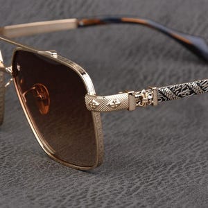 Puede incluir: Gafas de sol de aviador doradas con lentes degradadas marrones. Las monturas presentan detalles intrincados, incluyendo elementos decorativos en el puente y las patillas. Las patillas tienen un dise&ntilde;o negro y dorado.