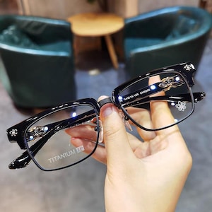 Op de afbeelding: Zwarte rechthoekige bril met decoratieve gouden accenten op de veren en het montuur. De glazen zijn helder en het montuur is voorzien van de tekst "TITANIUM IP". De bril wordt in een hand gehouden.