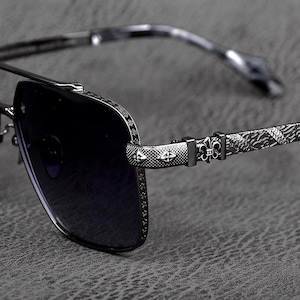 Puede incluir: Primer plano de gafas de sol con lentes oscuras y montura metálica decorativa. La montura presenta intrincados patrones y diseños, incluyendo símbolos de flor de lis y de cruz. Las patillas tienen un diseño texturizado y estampado. Las gafas de sol están sobre un fondo gris.