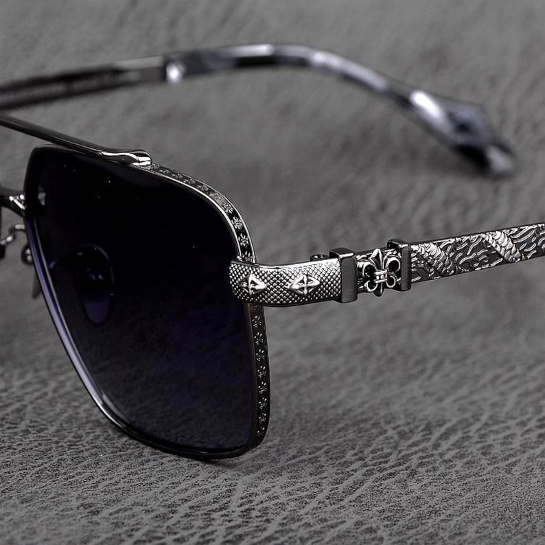 Puede incluir: Primer plano de gafas de sol con lentes oscuras y montura met&aacute;lica decorativa. La montura presenta intrincados patrones y dise&ntilde;os, incluyendo s&iacute;mbolos de flor de lis y de cruz. Las patillas tienen un dise&ntilde;o texturizado y estampado. Las gafas de sol est&aacute;n sobre un fondo gris.