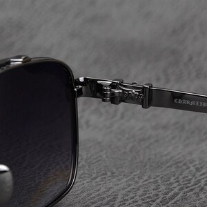 Puede incluir: Primer plano de gafas de sol negras con montura cuadrada. La bisagra y la patilla son de metal oscuro, con el texto "CHARMLIB" visible en la patilla. Las lentes son oscuras y el dise&ntilde;o general es elegante y moderno.