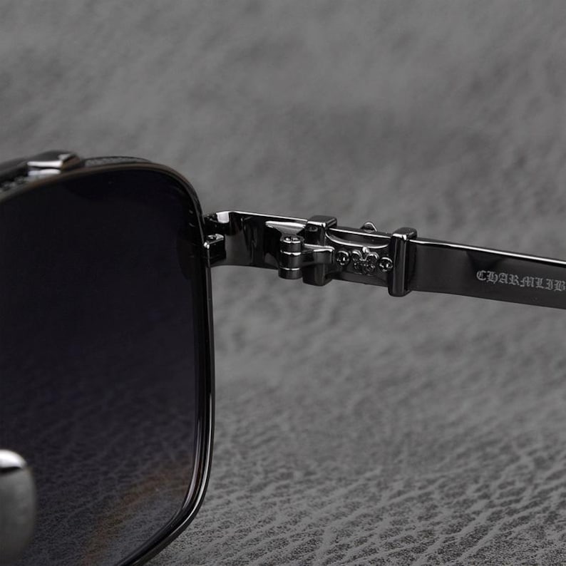 Puede incluir: Primer plano de gafas de sol negras con montura cuadrada. La bisagra y la patilla son de metal oscuro, con el texto "CHARMLIB" visible en la patilla. Las lentes son oscuras y el dise&ntilde;o general es elegante y moderno.