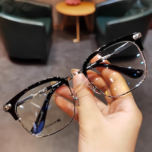 以下が含まれることがあります： 黒とシルバーの長方形フレームの眼鏡。 チタンフレームの眼鏡は手で持たれています。 レンズは透明で、テンプルは黒です。 モダンでスタイリッシュなデザインです。