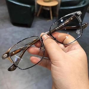 May include: Eyeglasses with a black metal frame and brown wooden arms. The glasses have a rectangular shape and are held by a hand. The arms have decorative metal accents. The lenses are clear.