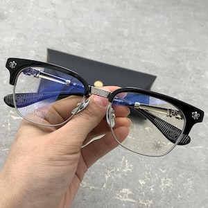 Op de afbeelding: Bril met een zwart bovenframe en zilveren onderranden. Het ontwerp omvat decoratieve bloemaccenten op de slapen. De lenzen zijn helder. De bril wordt tegen een vage achtergrond gehouden. Het woord "TITANIUM" is zichtbaar op het frame.