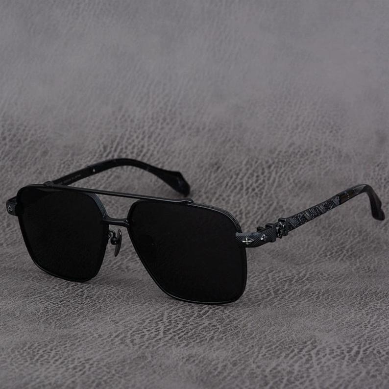 Puede incluir: Gafas de sol aviador negras con doble puente y lentes oscuras. Las monturas son de metal gris oscuro con patillas estampadas. Las gafas de sol est&aacute;n sobre un fondo gris.
