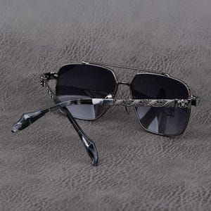 Puede incluir: Gafas de sol grises oscuras con doble puente y lentes cuadradas. Las monturas son de color met&aacute;lico oscuro con detalles decorativos en las patillas. Las lentes son de color gris oscuro.