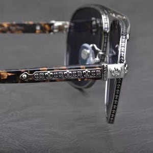 Puede incluir: Primer plano de gafas de sol con montura plateada y lentes oscuras. Las patillas tienen un patr&oacute;n de carey y est&aacute;n adornadas con detalles plateados decorativos. Las bisagras y los detalles de la montura presentan dise&ntilde;os intrincados.