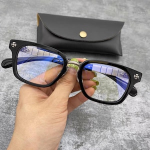 Op de afbeelding: Zwarte bril met vierkant montuur en een decoratief zilveren kruisje op elke poot. De bril heeft blauw getinte glazen en wordt getoond voor een zwart leren etui met een gouden knoopsluiting. Het montuur leest "TITANIUM FP".